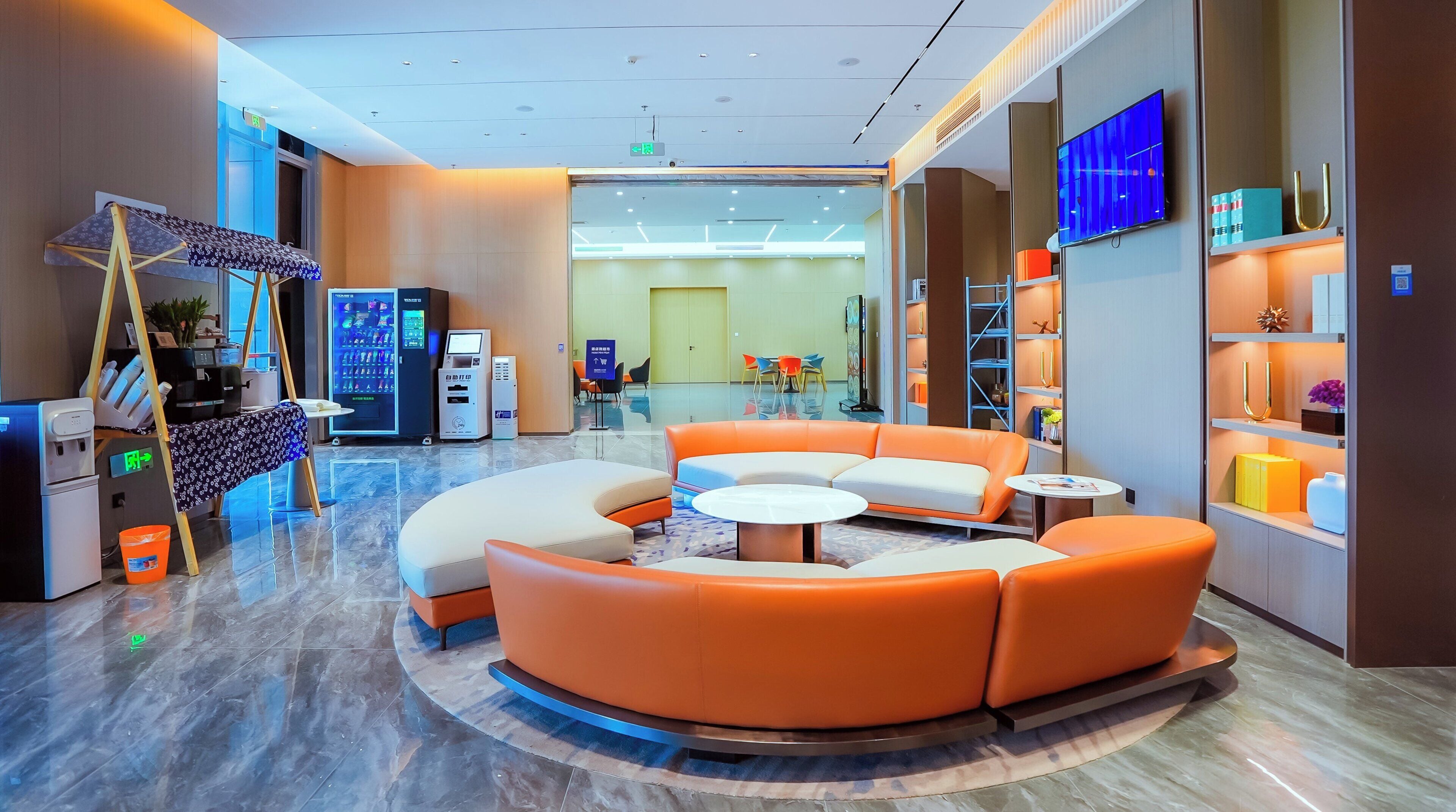 Un hall d’entrée moderne avec un canapé orange aux lignes courbes, une table basse ronde, des distributeurs automatiques et un téléviseur fixé au mur.