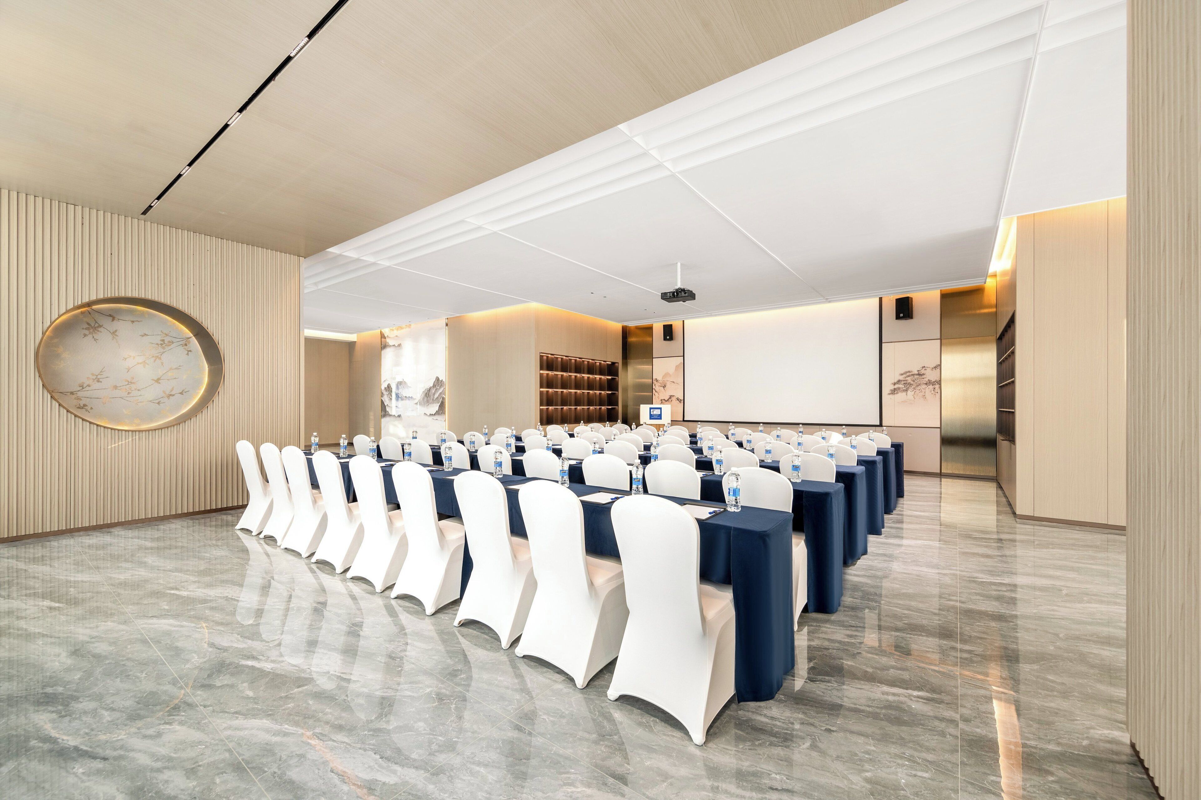 A conference room with rows of tables and chairs, a projector screen, and a large circular wall decoration.