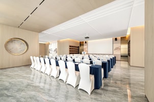 Un salón de conferencias con filas de mesas y sillas, una pantalla de proyector y un gran adorno circular en la pared.