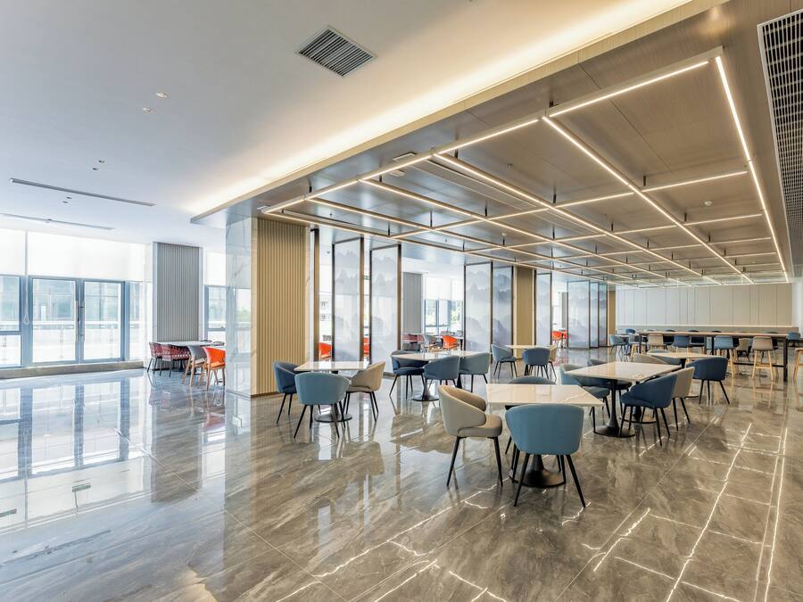 A spacious indoor area with tables and chairs, large windows, and a modern ceiling design.