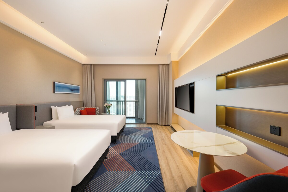 Habitación de hotel con dos camas, televisión, una mesita y vista a la ciudad.