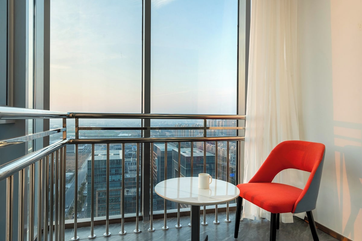 Un balcón con una silla roja y una pequeña mesa, con vistas a un paisaje urbano a través de amplias ventanas.