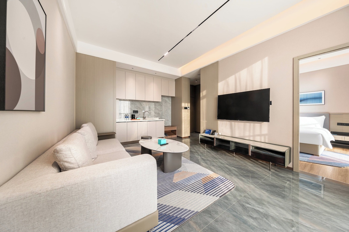 Una habitación de hotel moderna con sofá, una mesa de centro, televisor de pantalla plana, cama y una pequeña cocina.