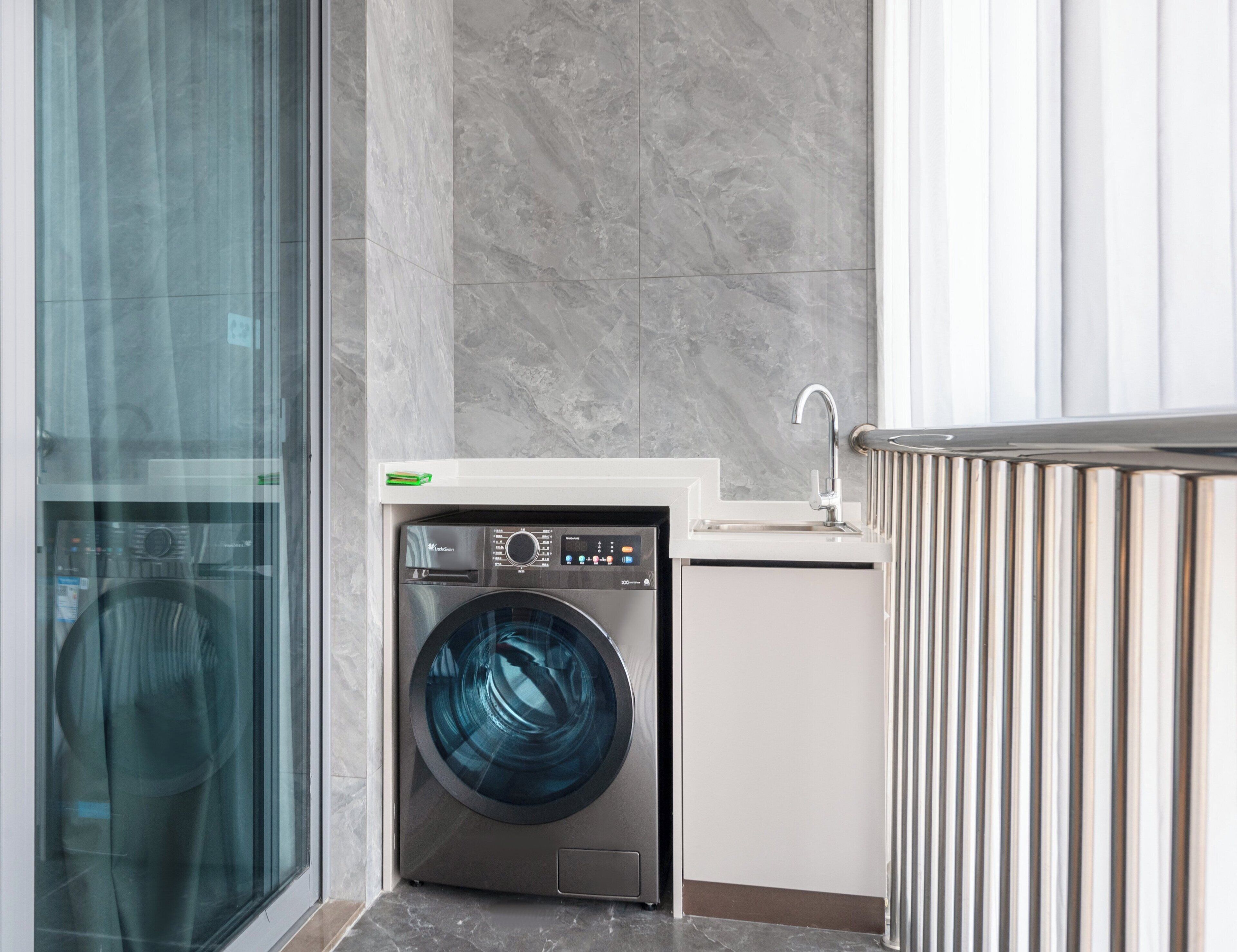 A compact laundry area with a front-loading washing machine, a small sink, and a glass door leading to a hallway.