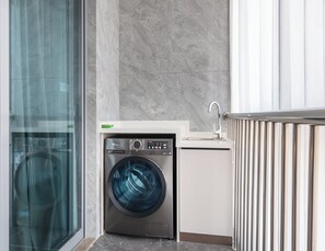 A compact laundry area with a front-loading washing machine, a small sink, and a glass door leading to a hallway. - Holiday Inn Express Qidong Economic Zone by IHG (Nantong)