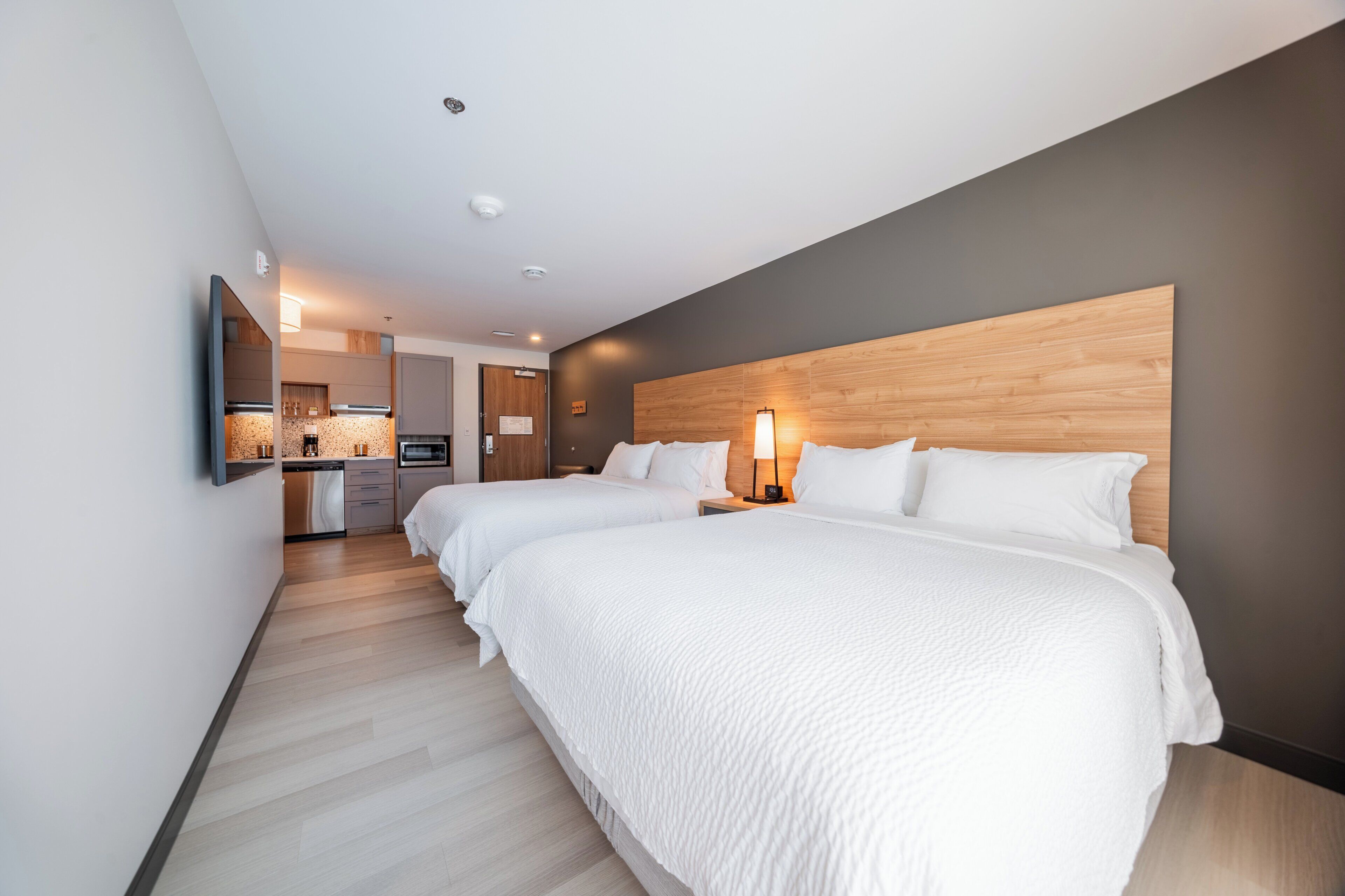 Une chambre d’hôtel moderne avec deux lits, une tête de lit en bois, un coin cuisine et un téléviseur à écran plat.