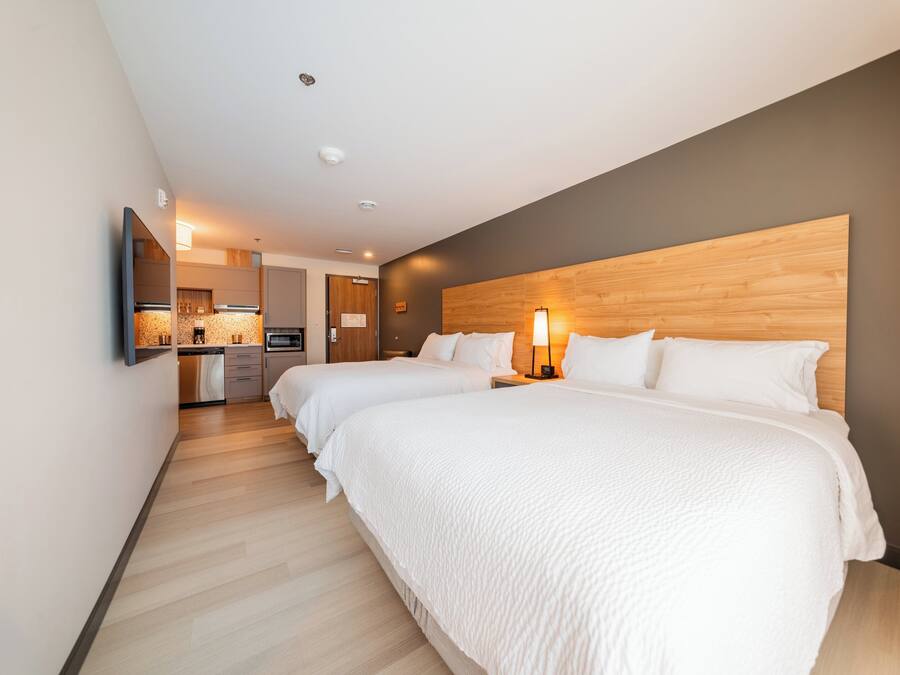 A hotel room with two beds, a wooden headboard, a kitchen area, and a flat-screen TV mounted on the wall.