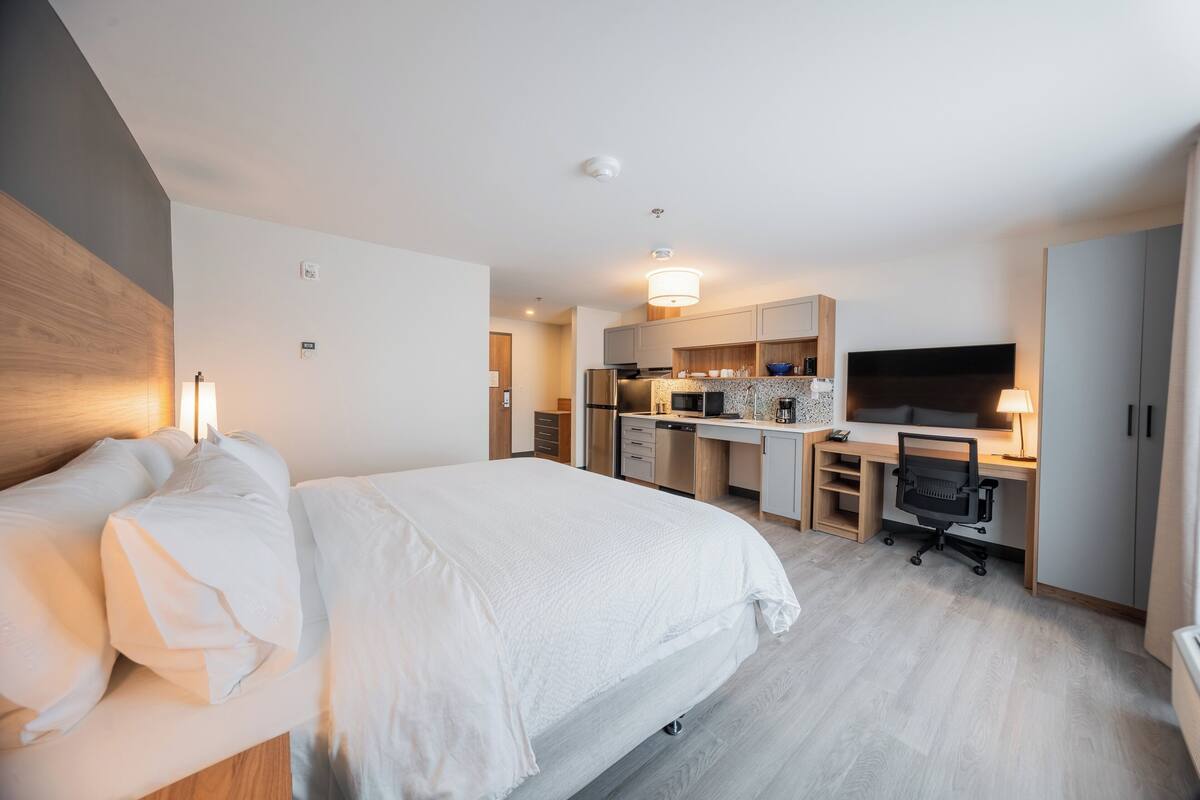 Habitación de hotel moderna con una cama grande, kitchenette, escritorio y televisor.