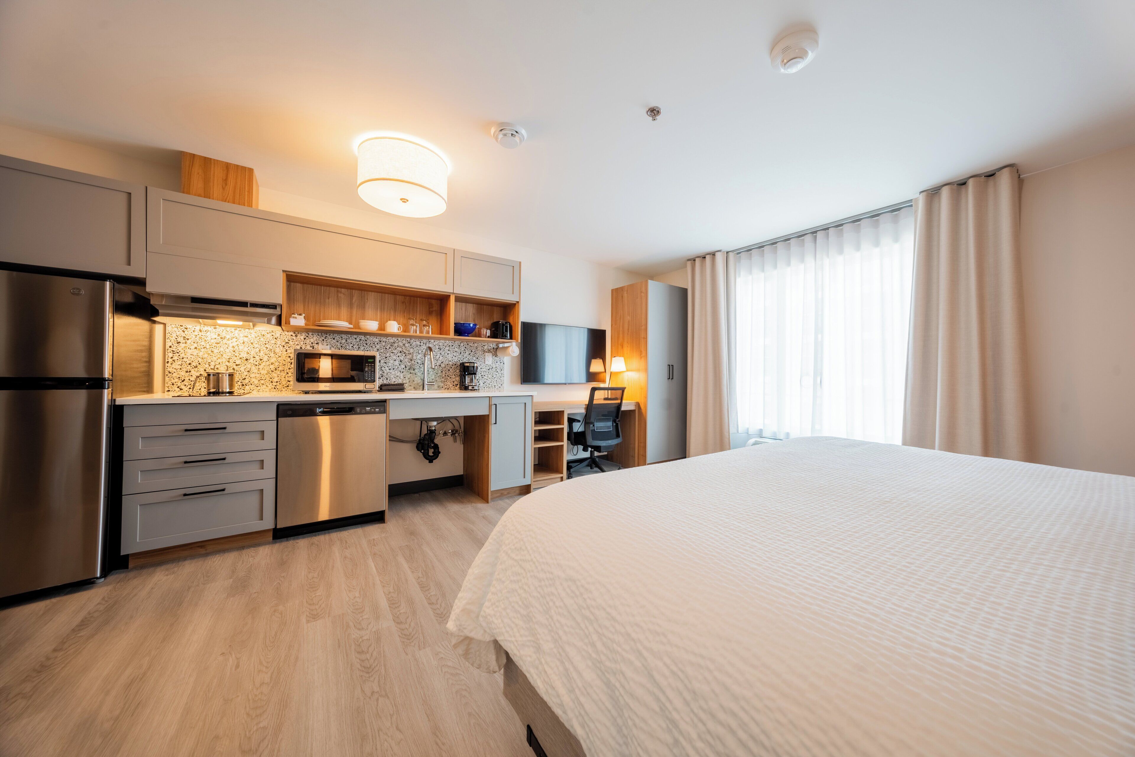 Ett modernt hotellrum med ett litet kök, en säng, ett skrivbord med en stol och ett stort fönster med gardiner.
