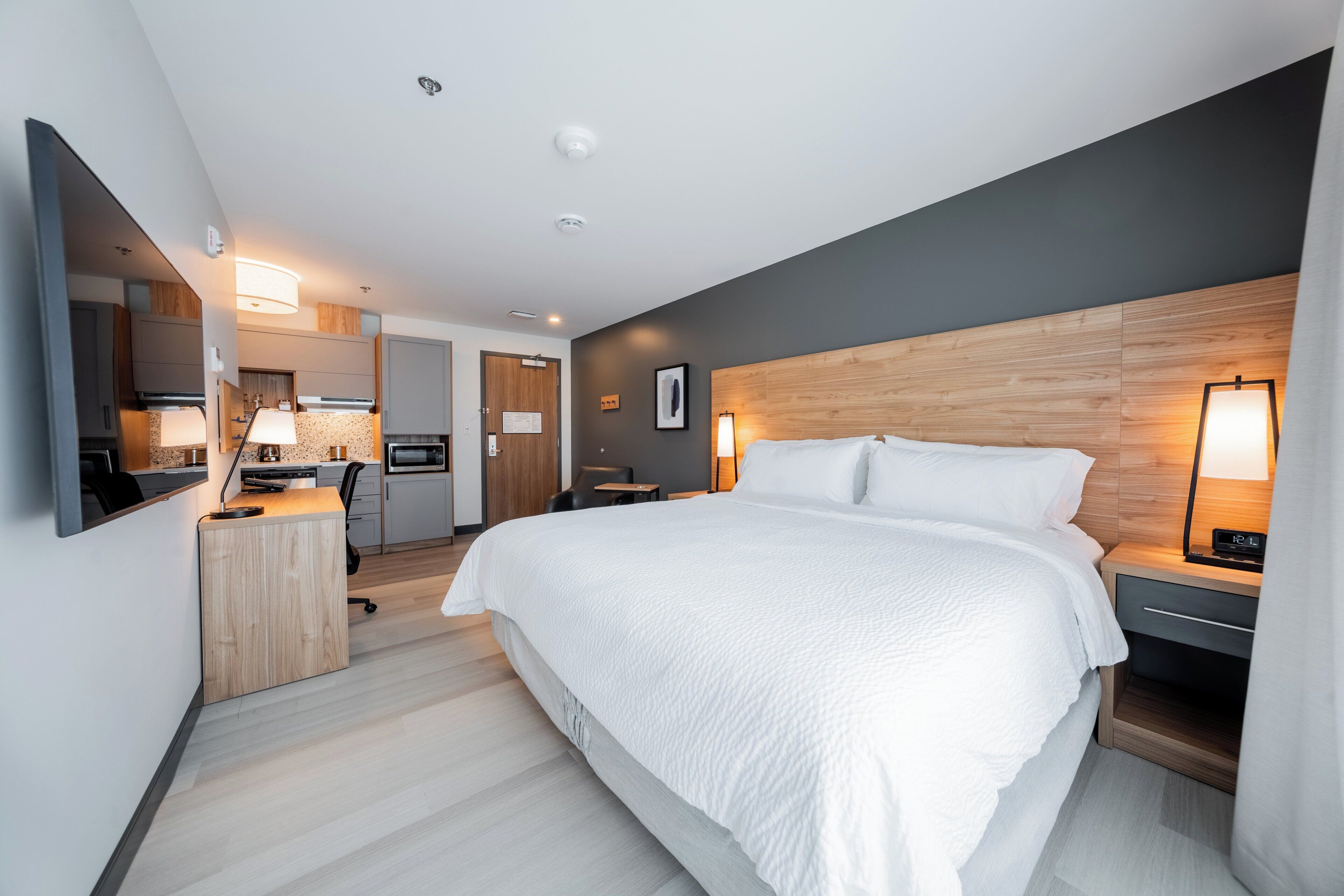 A modern hotel room with a large bed, a desk with a lamp, a microwave, and a small table.