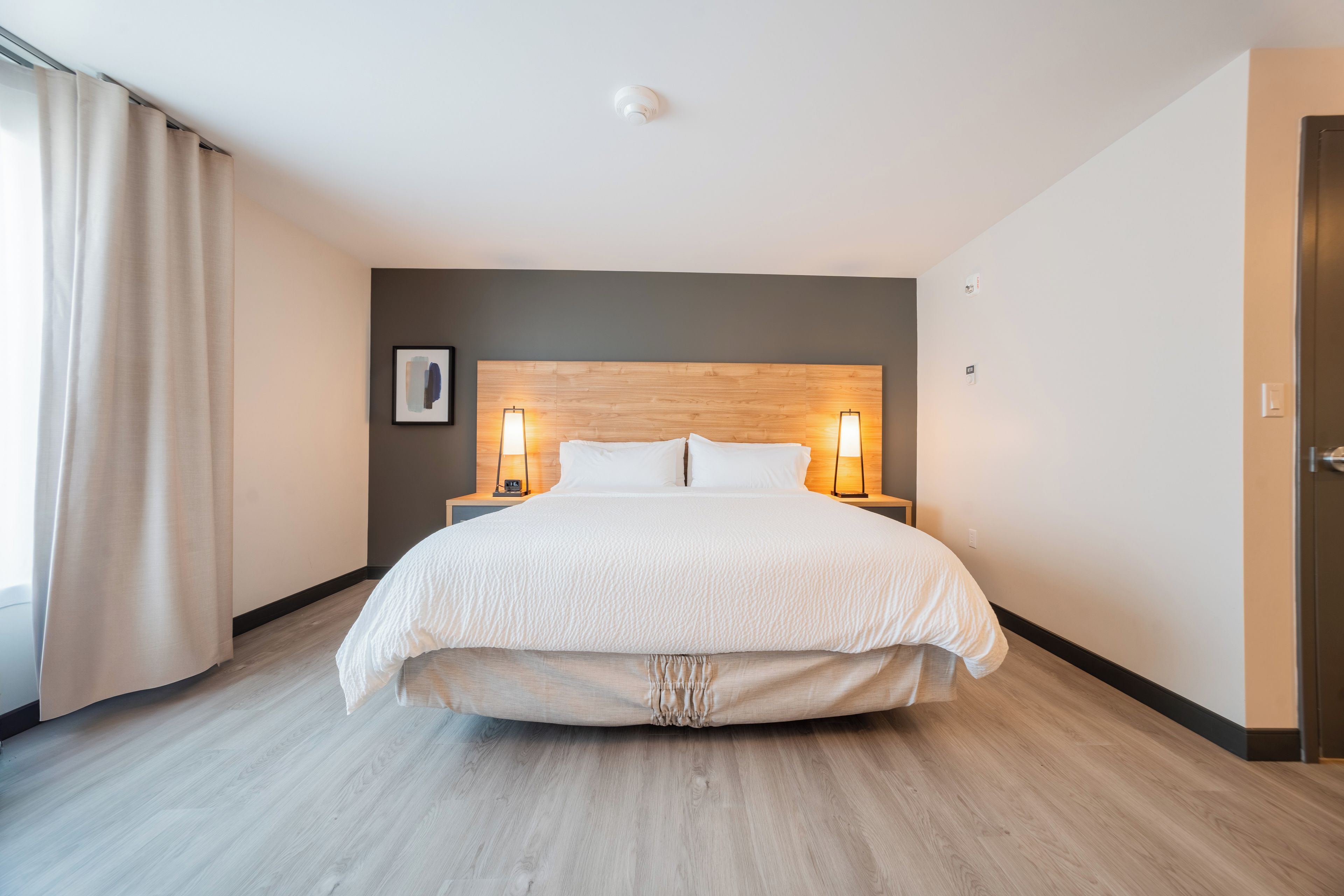 A modern hotel room with a large bed, two bedside lamps, a wooden headboard, and a view of the city through the window.