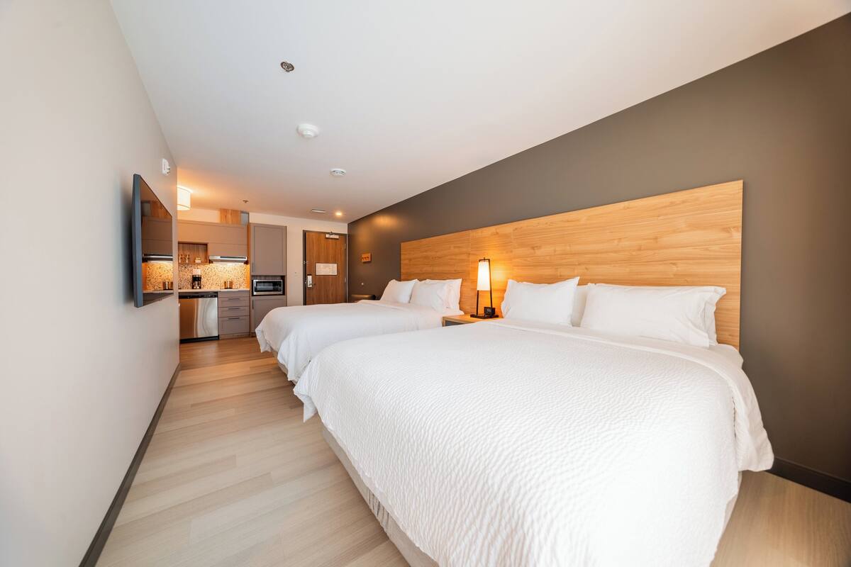 Una habitación de hotel con dos camas, cabecera de madera, zona de cocina y televisor de pantalla plana montado en la pared.
