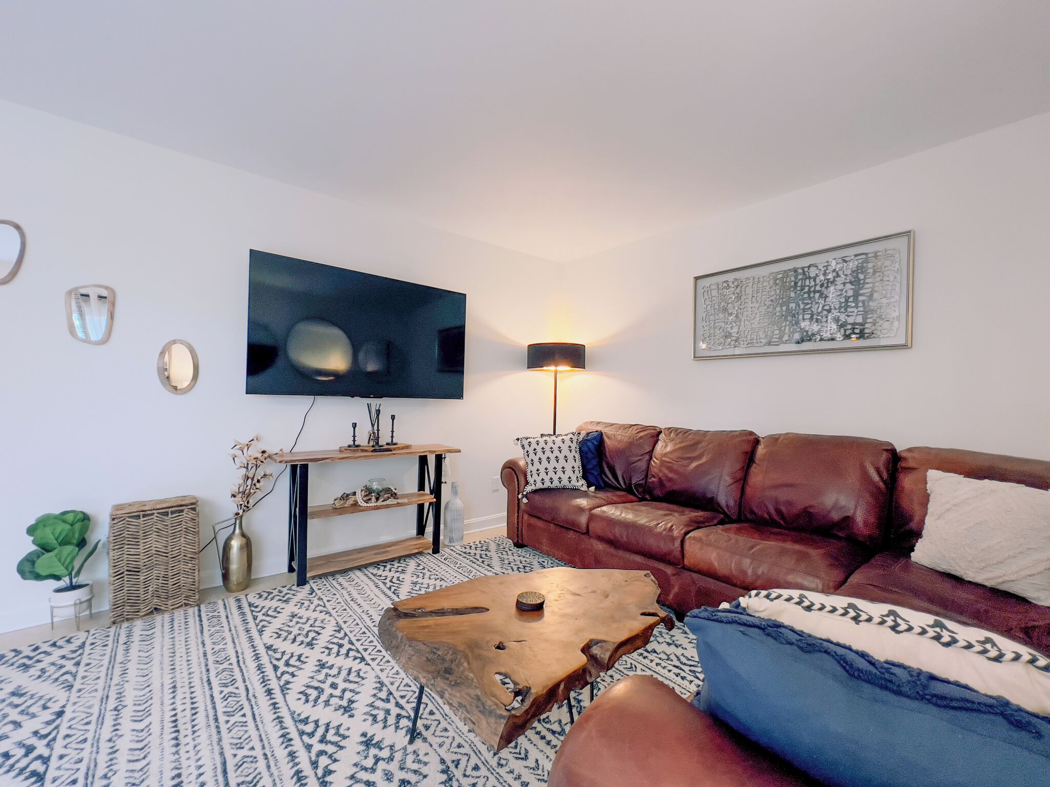 A living room with a brown leather sofa, a wooden coffee table, a patterned rug, a flat-screen TV, a floor lamp, and a wall-mounted shelf with decorative items.