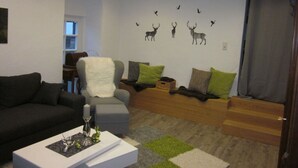 A living room with a grey sofa, a white armchair, and a coffee table. There is a wall with a mural of deer and birds.