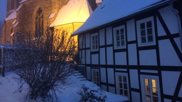A traditional half-timbered house with a snow-covered roof and a church in the background.