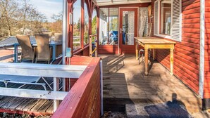 Une véranda couverte avec une table et des chaises en bois, une porte vitrée donnant sur l’intérieur et une vue sur les environs.