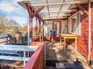 Une véranda couverte avec une table et des chaises en bois, une porte vitrée donnant sur l’intérieur et une vue sur les environs.
