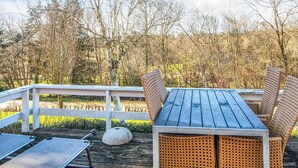 Une terrasse en bois, agrémentée d’une table et de chaises, entourée d’une balustrade blanche, et offrant une vue sur une zone boisée.