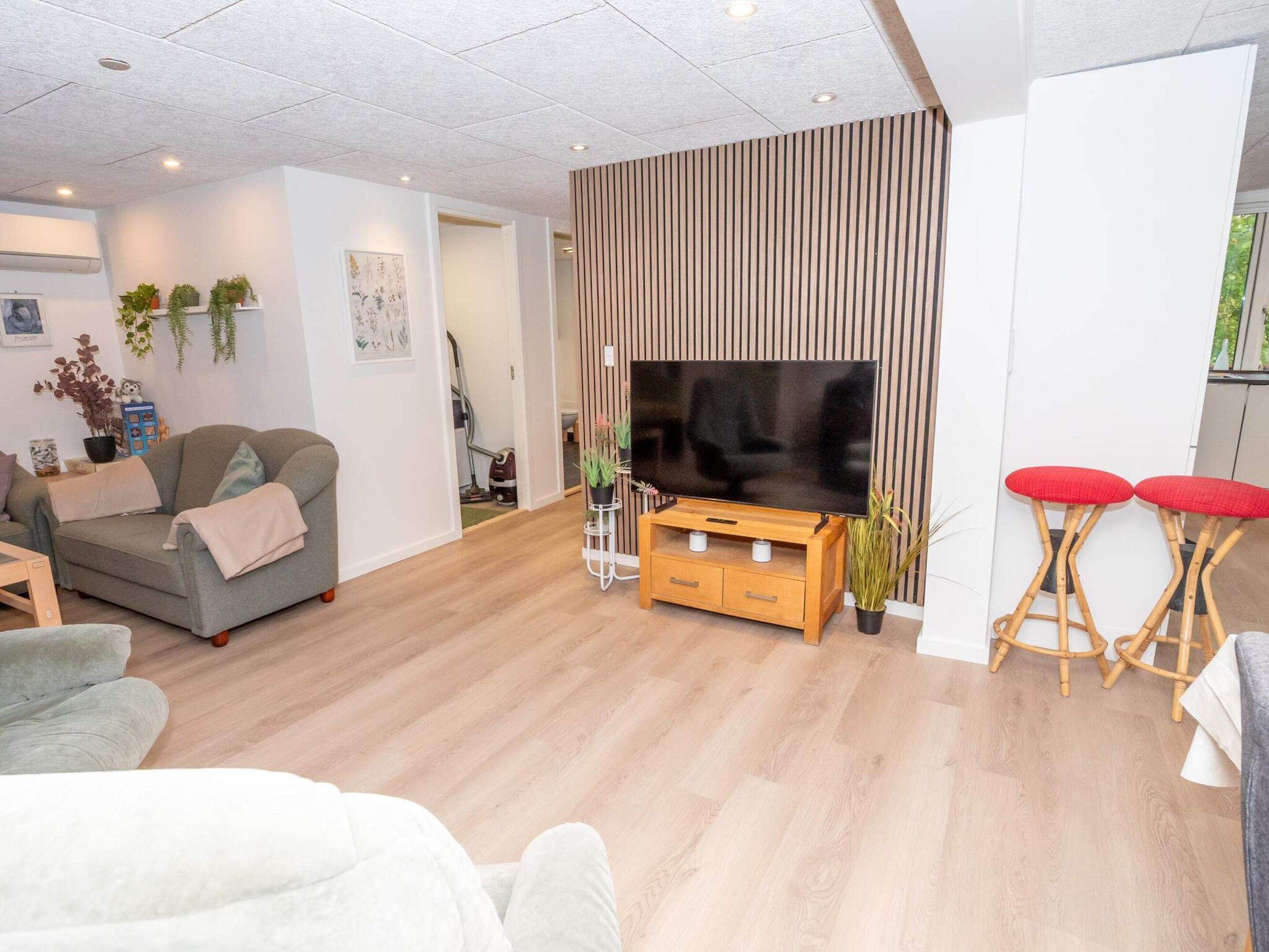A modern living room with a flat-screen TV, wooden flooring, a sofa, a coffee table, and two red bar stools.