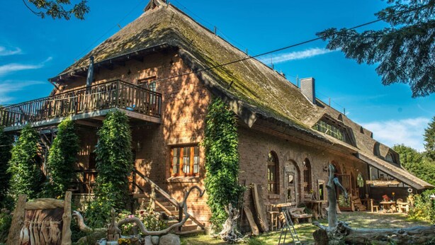 A rustic building with a thatched roof, a balcony, and a garden area with wooden furniture and plants. - Quellenhof Comfortable Holiday Residence (Retschow)