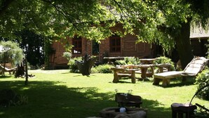 A rustic outdoor seating area with wooden furniture and a stone table, surrounded by lush greenery and a brick building in the background. - Quellenhof Comfortable Holiday Residence (Retschow)
