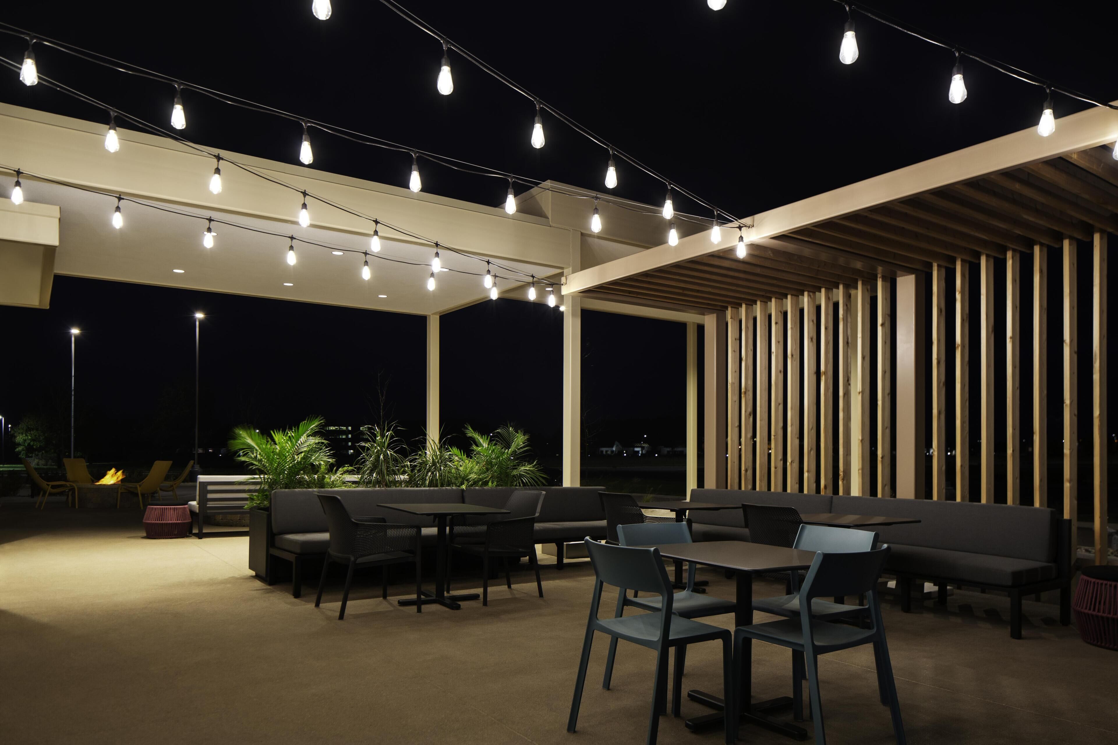 An outdoor patio area with tables and chairs, string lights overhead, and a wooden slatted wall.