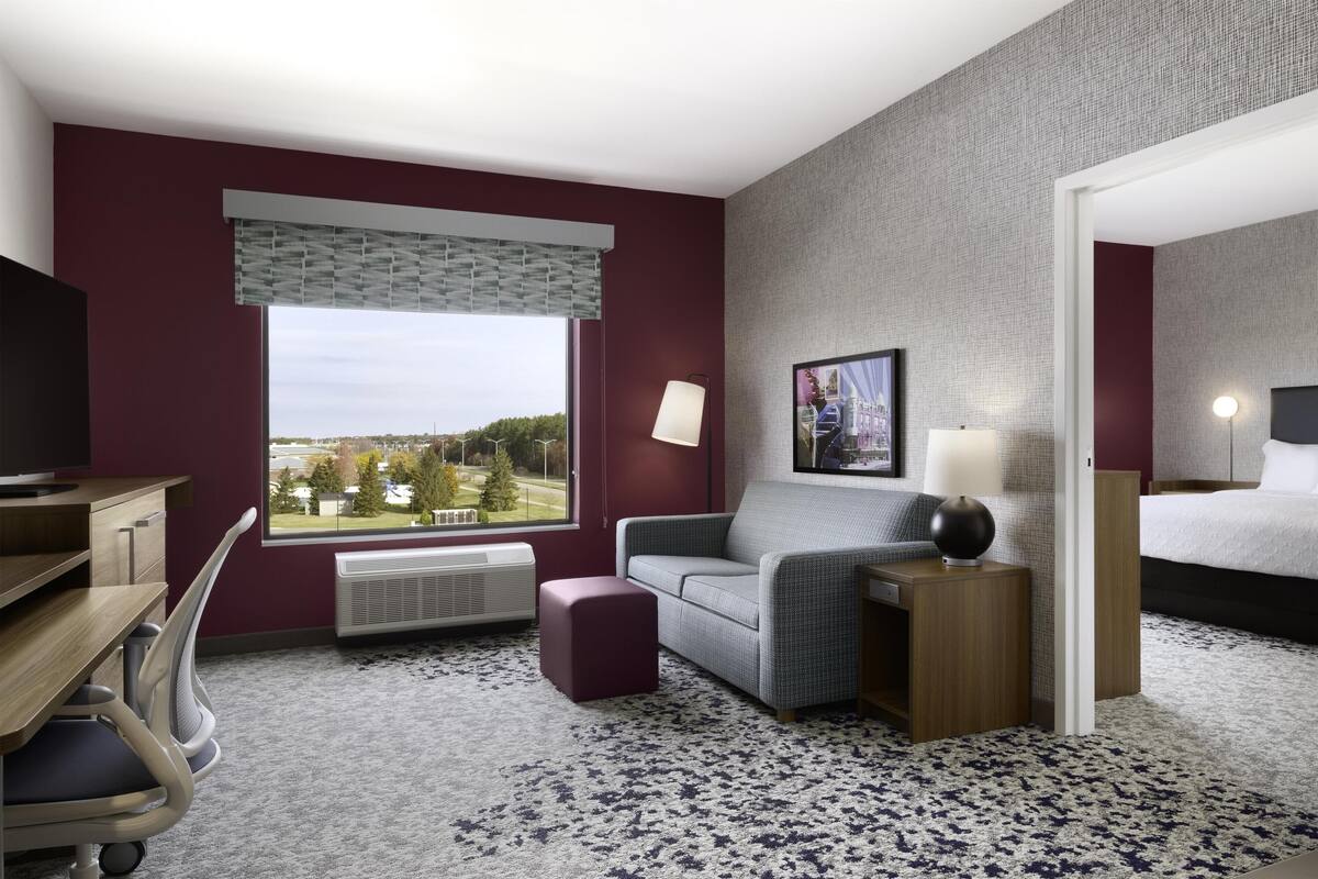 A hotel room with a desk, a cushioned seating area, a bed, a large window, and a television.