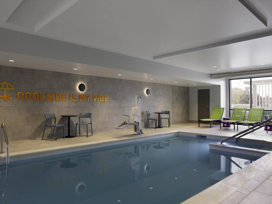 Un área de piscina interior moderna con vista despejada a la piscina, sillas de descanso y una pared con el texto 'POOLSIDE IS MY VIBE'.