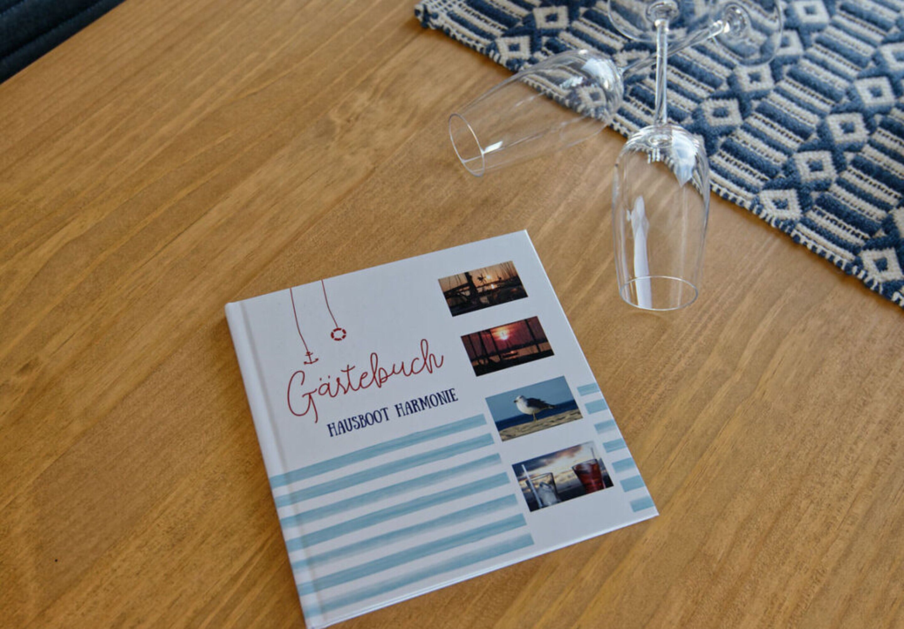 A book titled "Gartebuch Hausboot Harmonie" on a wooden table with two empty wine glasses and a patterned rug.