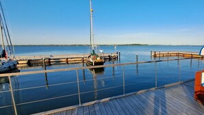 A wooden deck with a railing overlooking calm water, a dock with a sailboat, and a distant shoreline. - Houseboat Harmony (Stralsund)