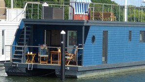 A blue houseboat with a covered deck area, a small outdoor seating area, and a staircase leading to an upper deck. - Houseboat Harmony (Stralsund)