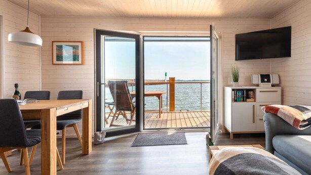 A modern interior with a wooden floor, a dining area with a table and chairs, a sofa, a TV, and a view of the sea through a glass door. - Houseboat Harmony (Stralsund)
