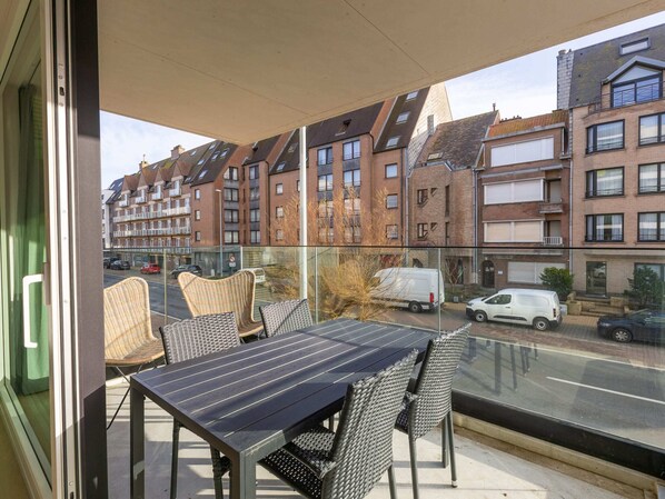 Un balcón con sillas de mimbre y una mesa, con vistas a una calle con coches aparcados y edificios residenciales.