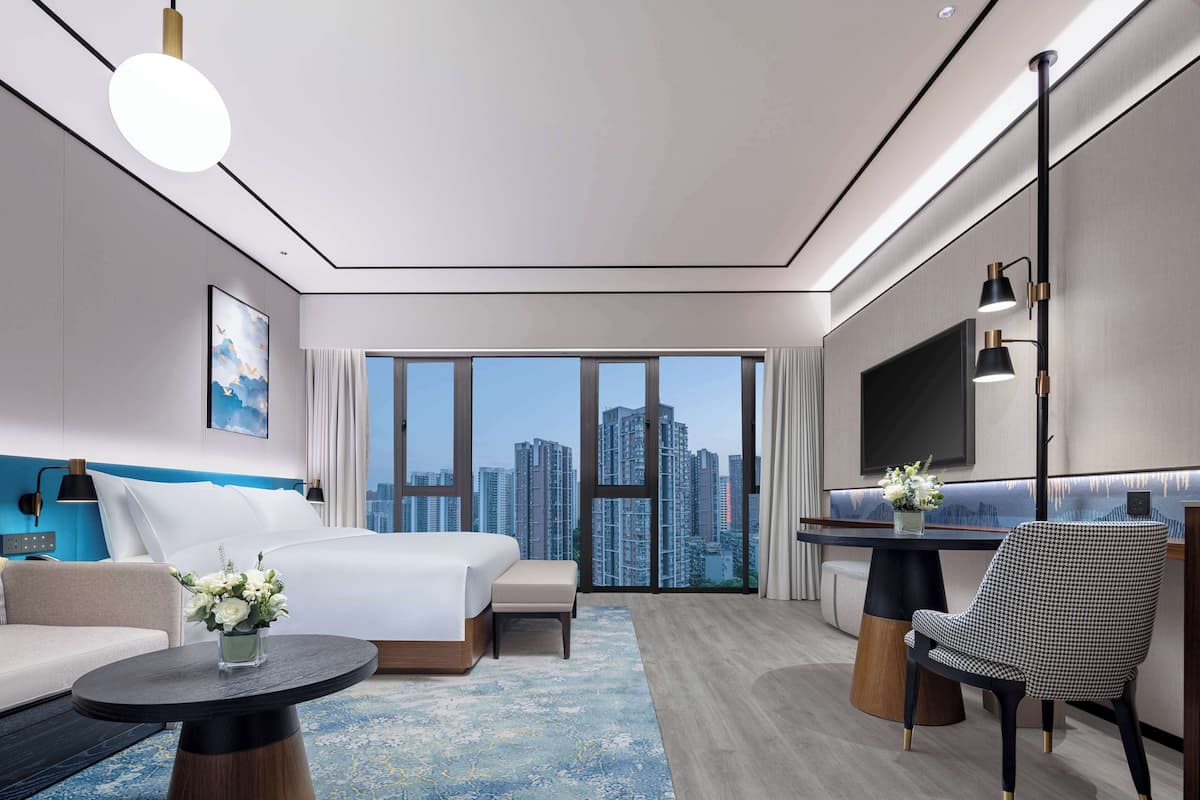 A modern hotel room with a large bed, a sofa, a dining table, and a TV. The room has a city view through large windows.