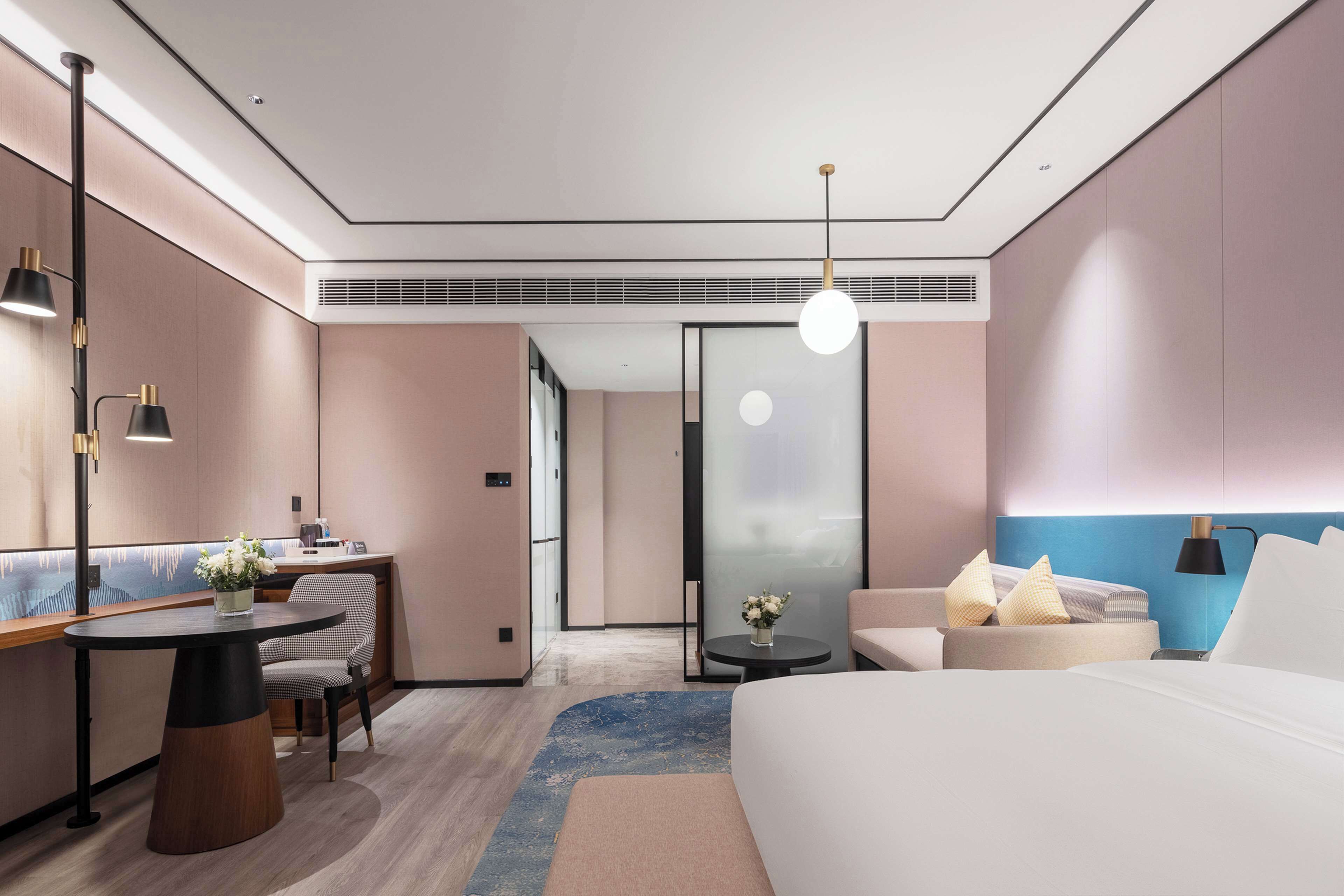 A modern hotel room with a bed, a desk, a chair, a round table, and a small side table with a vase of flowers.