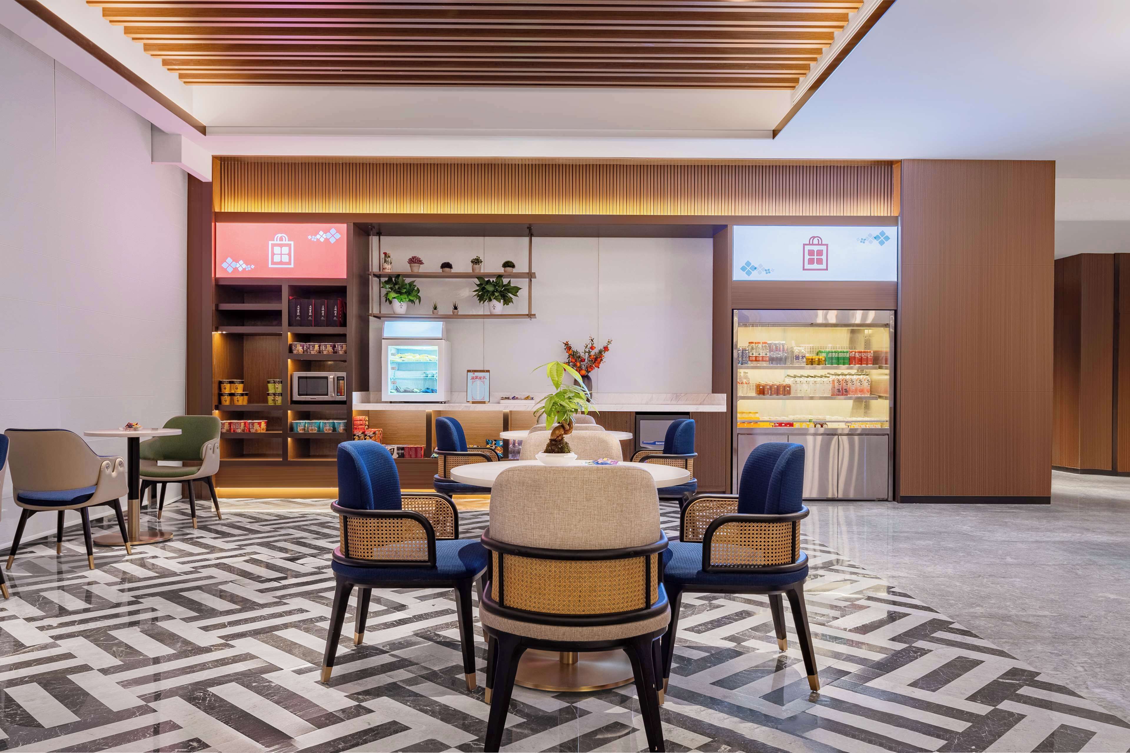 Un hall d’hôtel moderne avec une table ronde, des chaises bleues, un coin café et une bibliothèque agrémentée d’objets décoratifs.