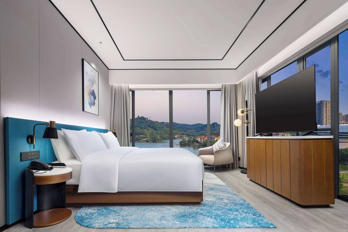 A modern hotel room with a large bed, a TV, and a view of a cityscape.