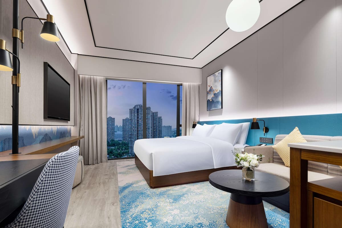 A modern hotel room with a large bed, a desk, a sofa, and a view of a cityscape.