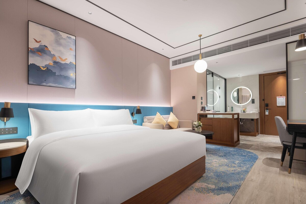 A modern hotel room with a large bed, a desk, a round mirror, and a bathroom with a vanity.