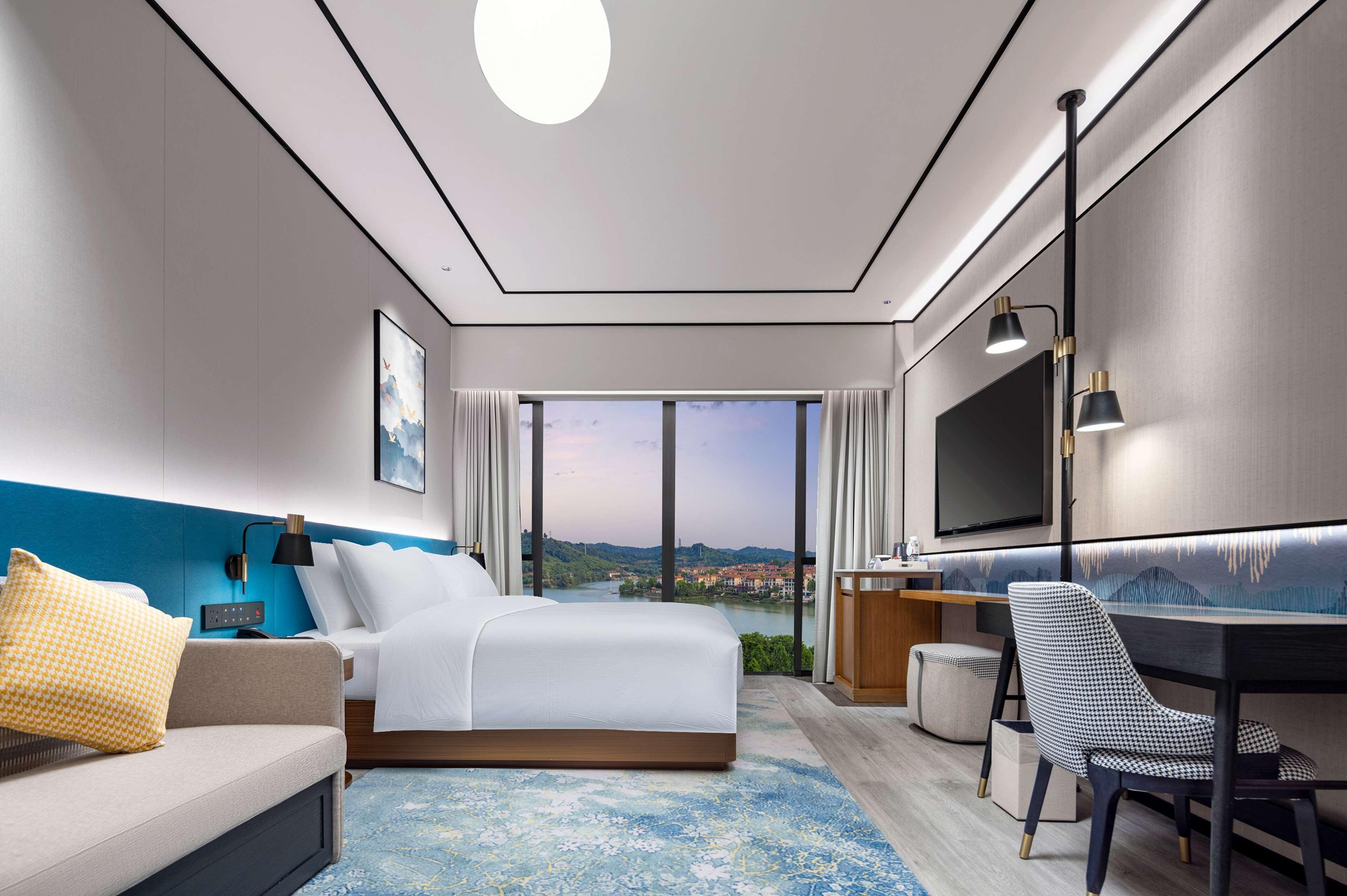 Moderni hotellihuone, jossa on suuri sänky, työpöytä, tuoli, televisio ja näkymä kaupunkimaisemaan.