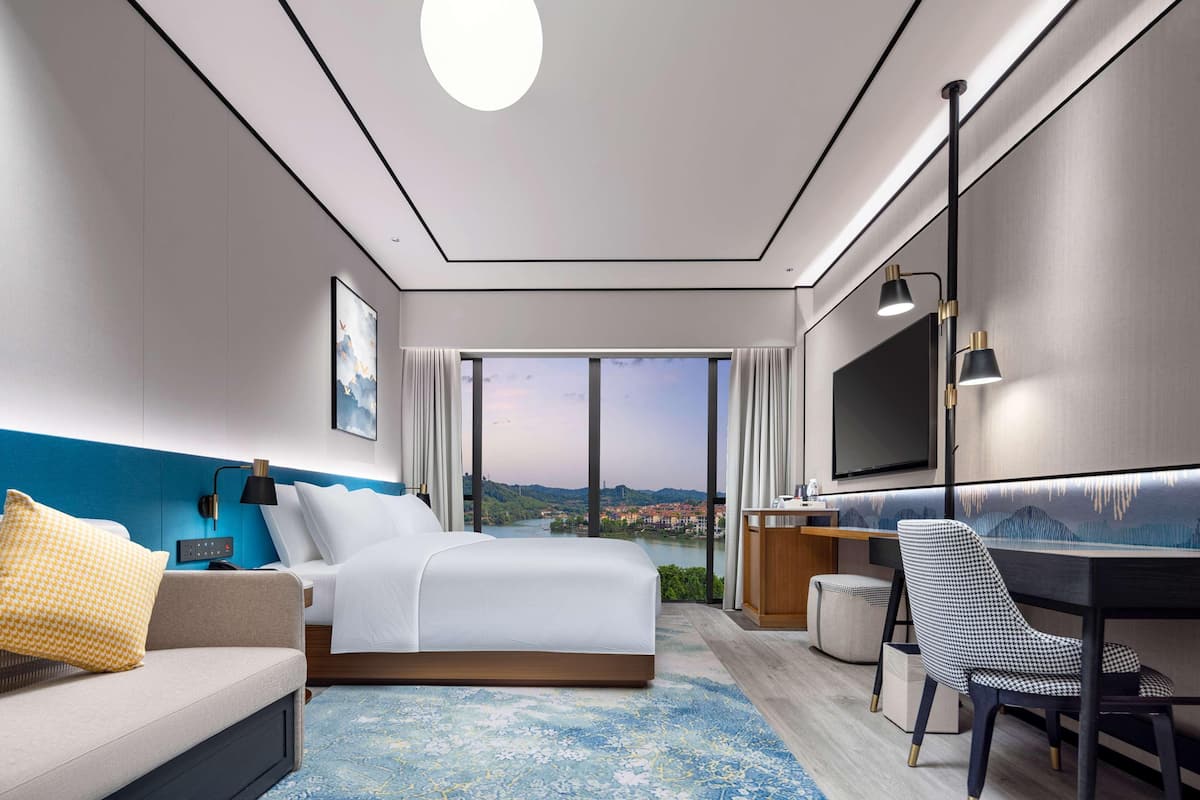A modern hotel room with a large bed, a desk, a chair, a TV, and a view of a cityscape.