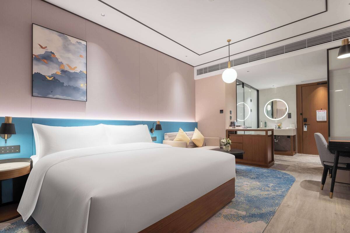 A modern hotel room with a large bed, a desk, a chair, a round mirror, and a bathroom with a sink and mirror.