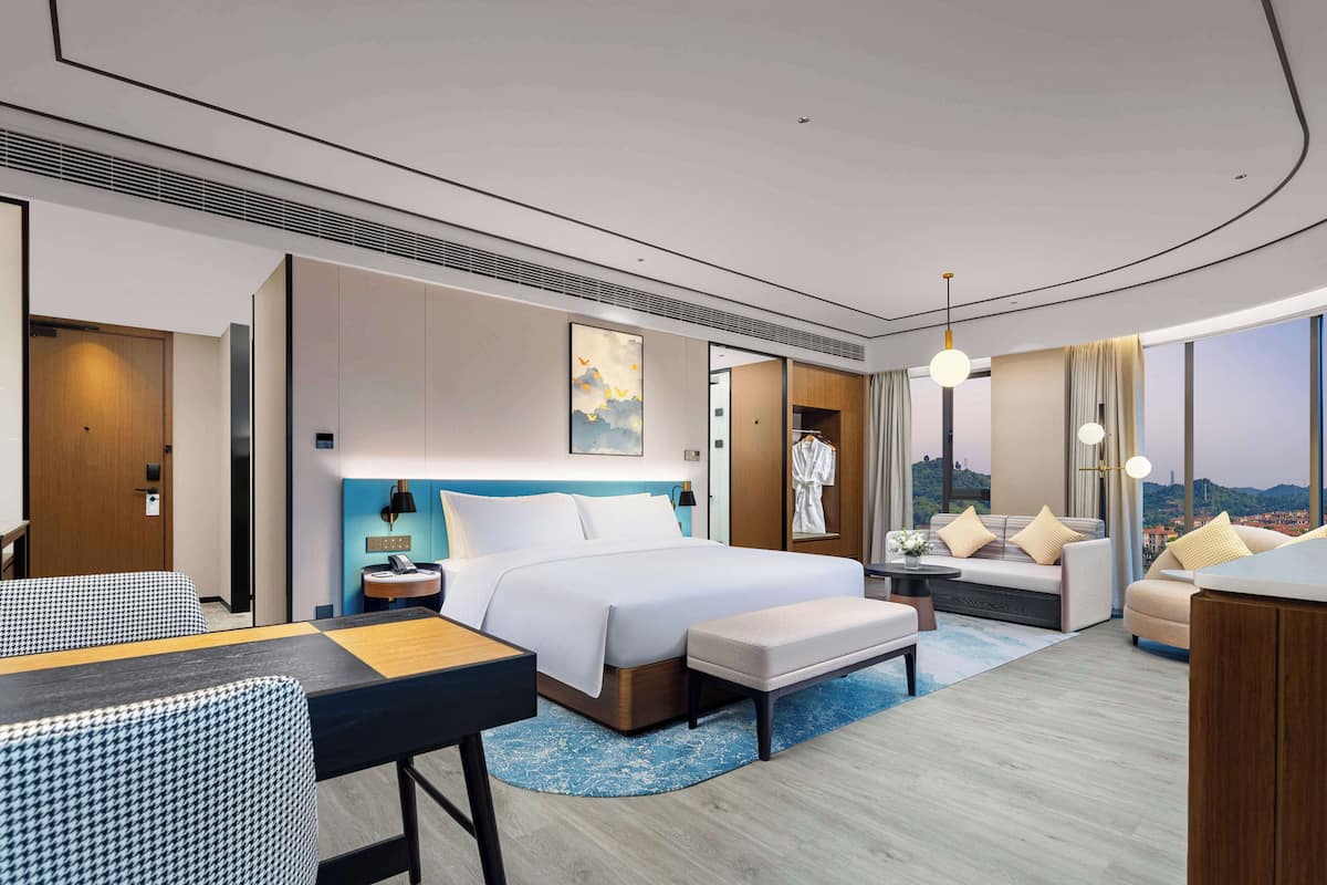 A modern hotel room with a large bed, a sofa, a desk, and a view of the city.