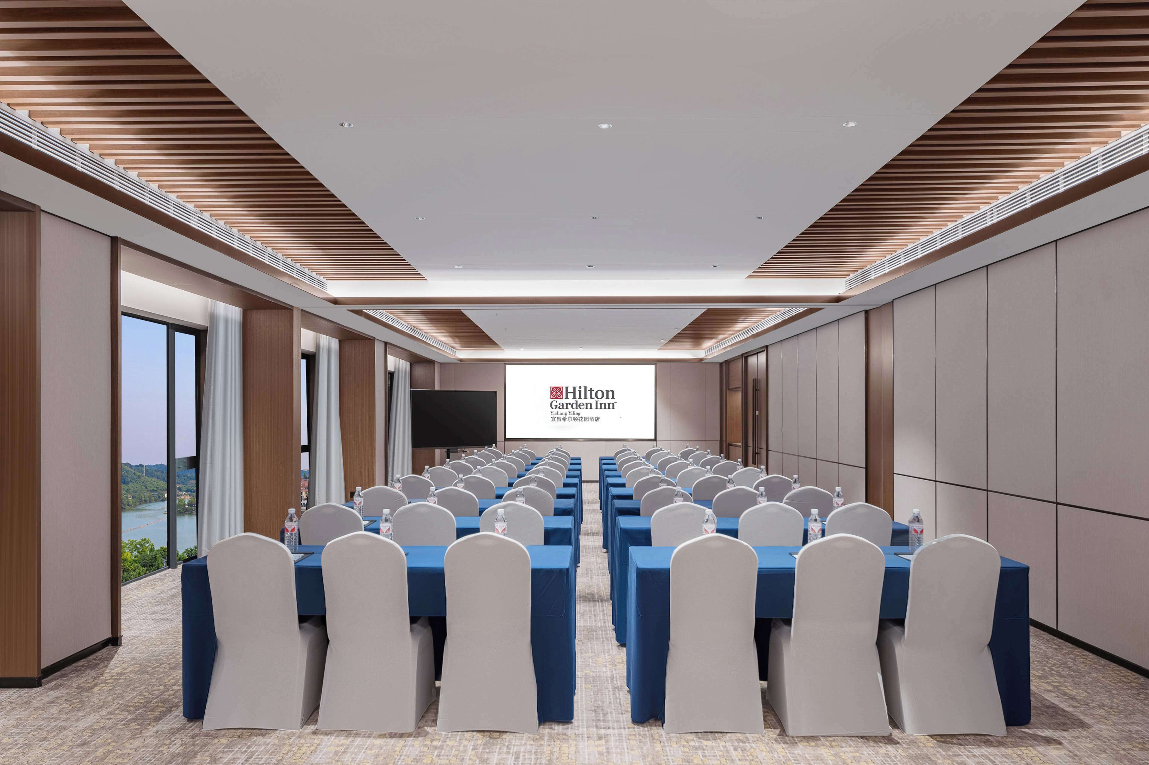 A conference room with rows of tables and chairs, a large screen displaying the Hilton Garden Inn logo, and a view of a river through large windows.