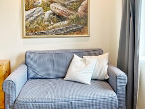 Een blauw geruit bankje met een kussen, een schilderij van rotsen en gras aan de muur, en een raam met gordijnen.
