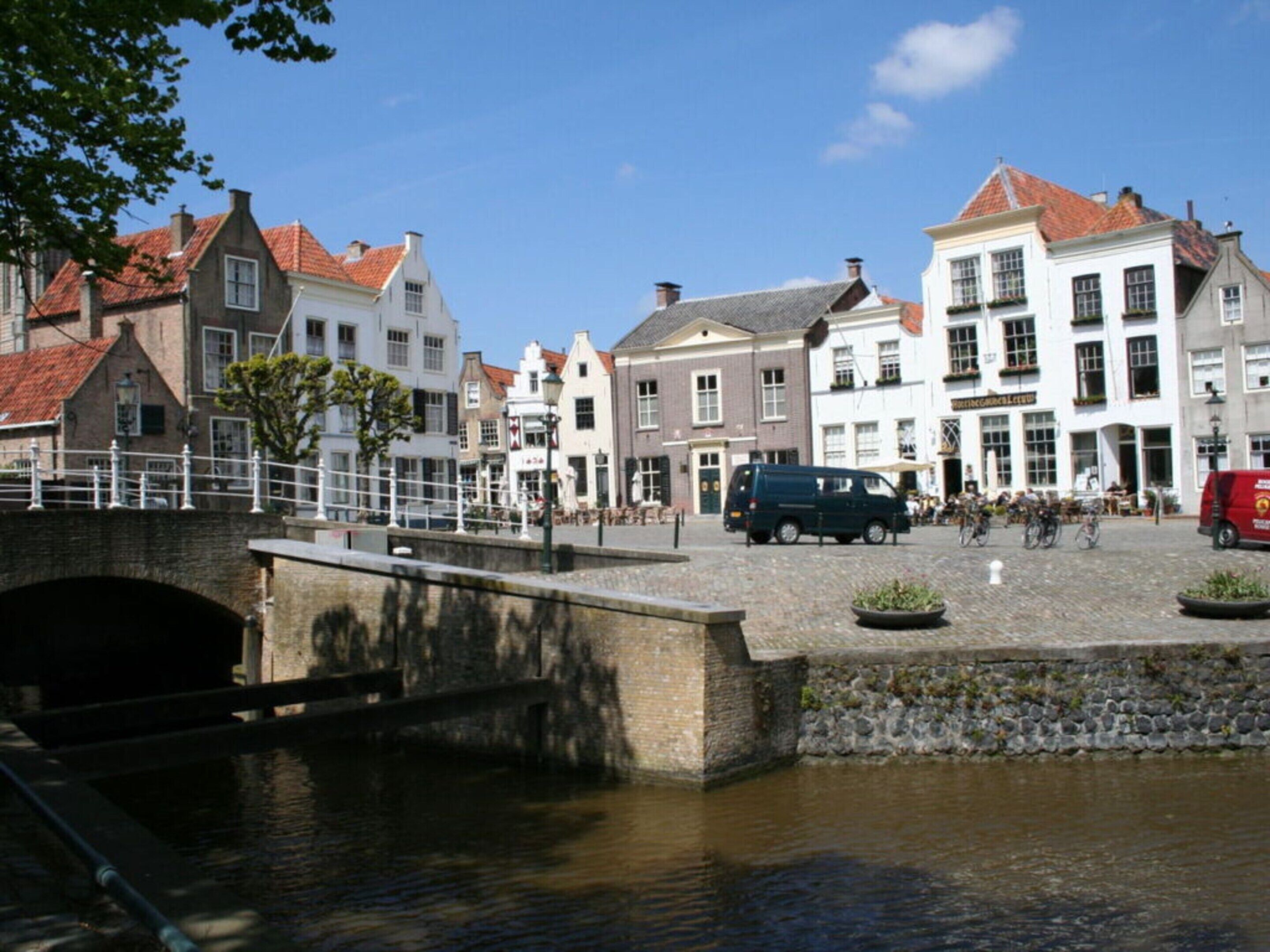 Uma rua à beira de um canal, com edifícios históricos, uma ponte e uma fileira de veículos estacionados.