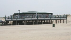 Un edificio frente al mar con techo verde y un piso superior de vidrio, probablemente un restaurante o cafetería, con una playa arenosa al fondo.