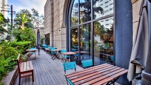 An outdoor patio with wooden tables and blue chairs, surrounded by greenery and a building with large windows. - HOTEL FARIA LIMA, LUXUOSO ALTO PADRÃO (São Paulo)