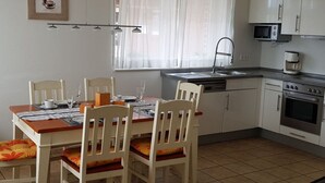 Une cuisine compacte avec des meubles blancs, un four à micro-ondes, un four traditionnel et un évier. On y trouve une table à manger avec des chaises et une fenêtre ornée de rideaux légers.