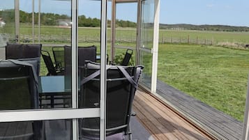 Un espacio de estar al aire libre techado, con paredes de vidrio, un piso de madera y vistas a un campo verde.
